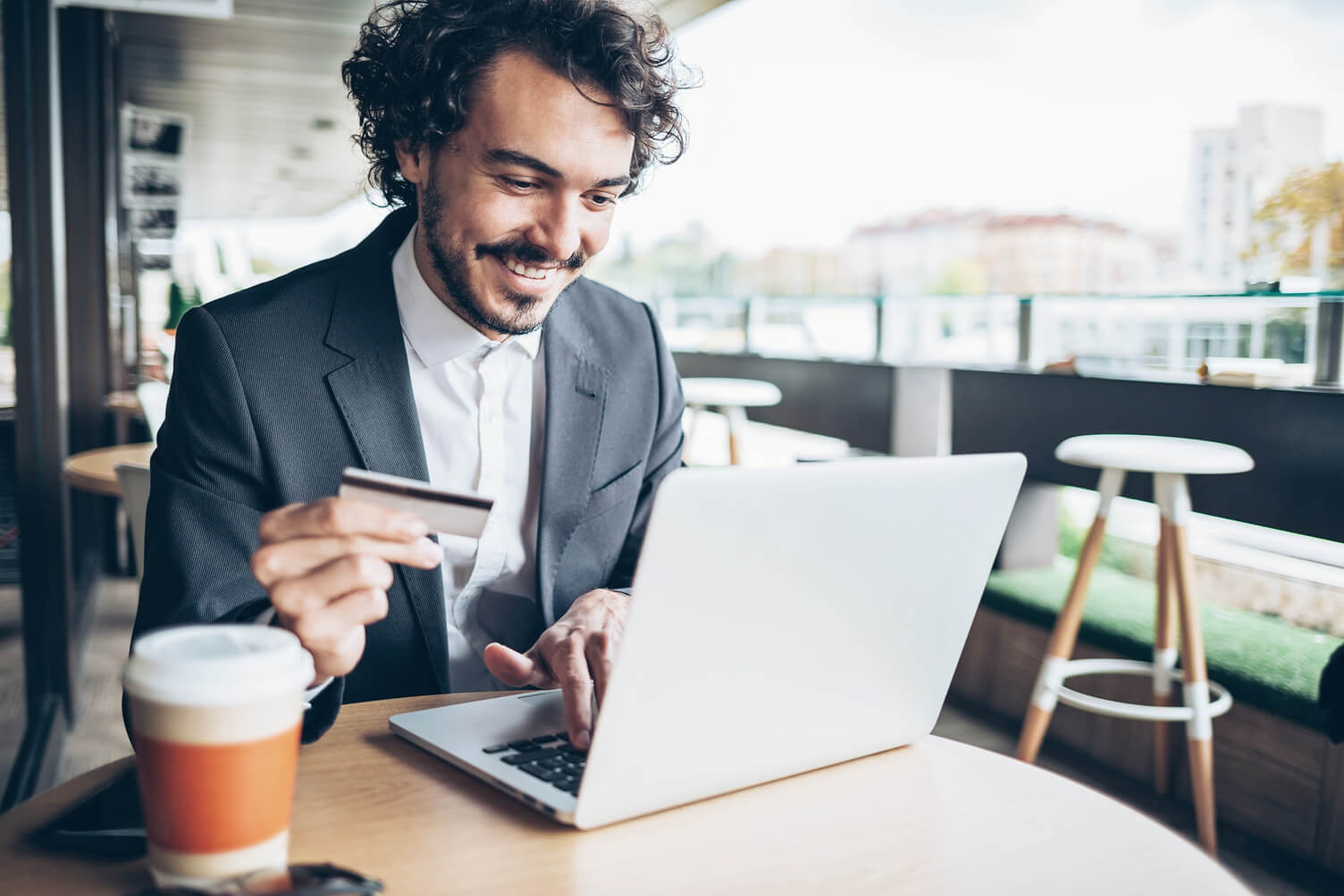 a man at a coffee shop holidng a credit card in one hand and looking at his laptop