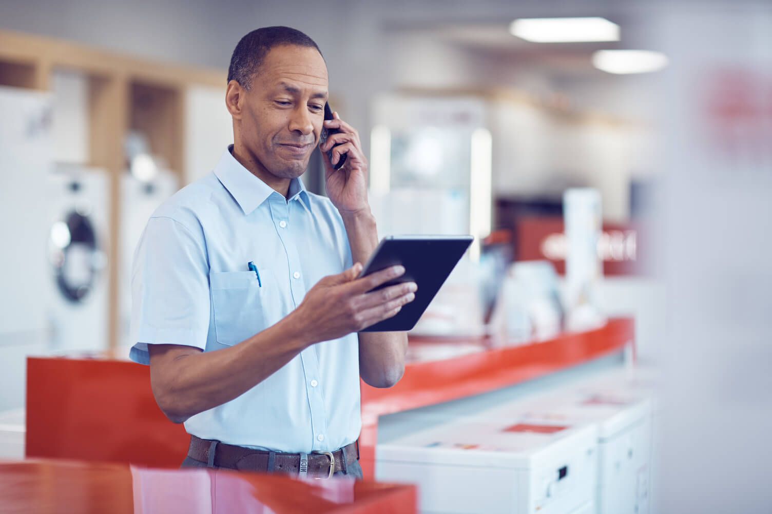 a man in a blue shirt holding a tablet in one hand and a phone up to his ear