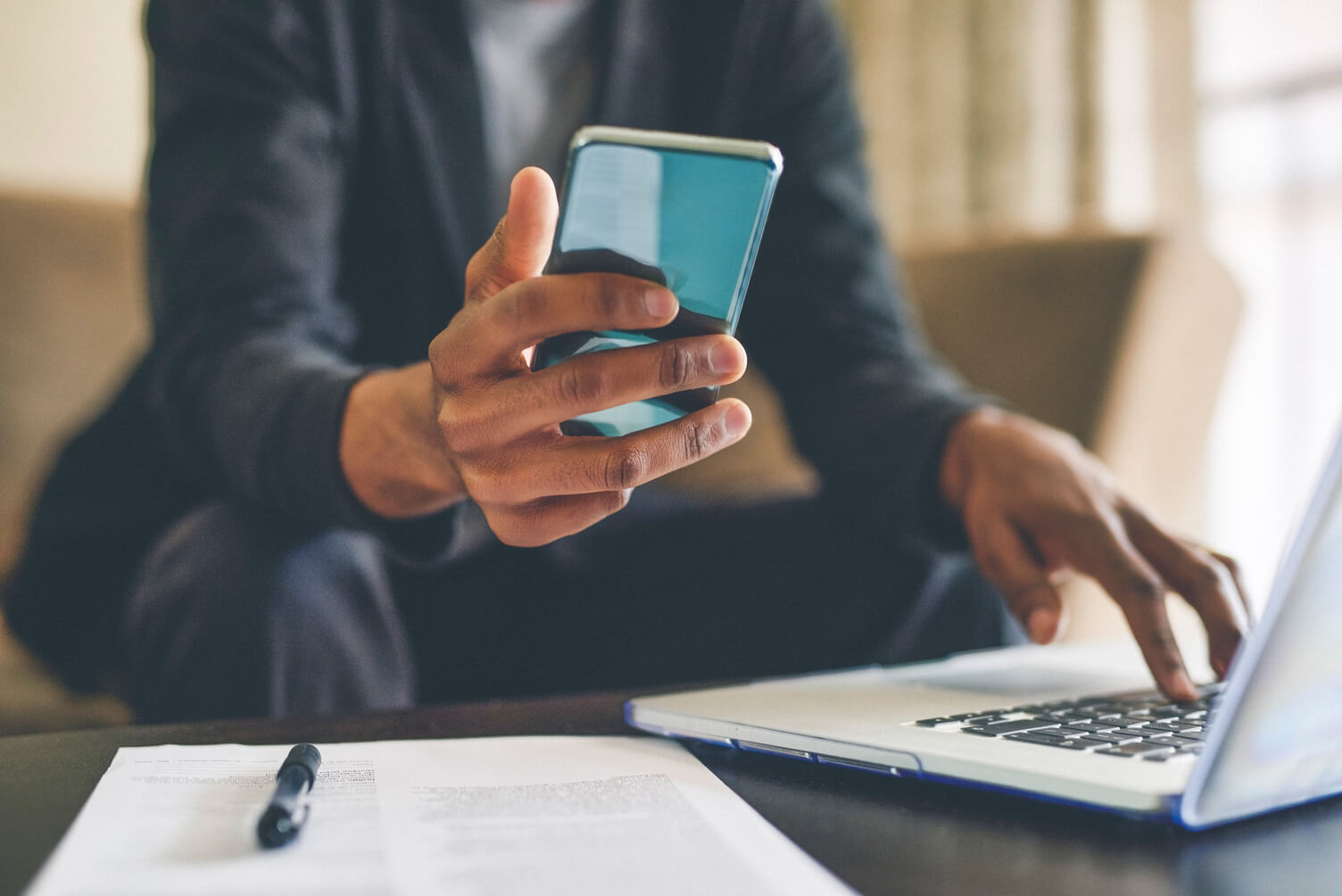 a cropped image of a mans hands, one hand is holding a phone the other typing on his laptop